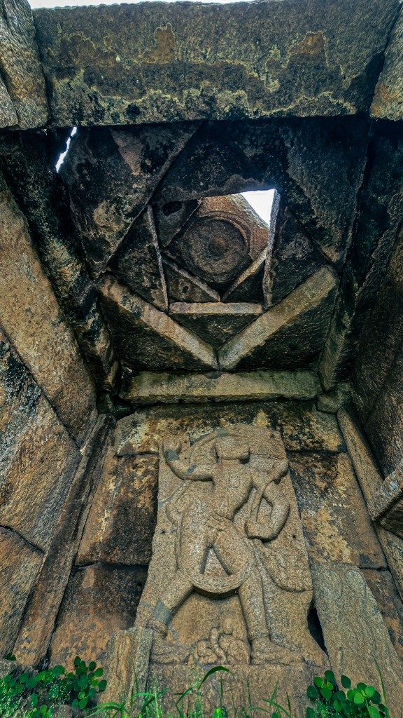 Close view inside a Hampi stone chamber — the star ceiling fills the frame above, the deity carving barely visible in the shadows below