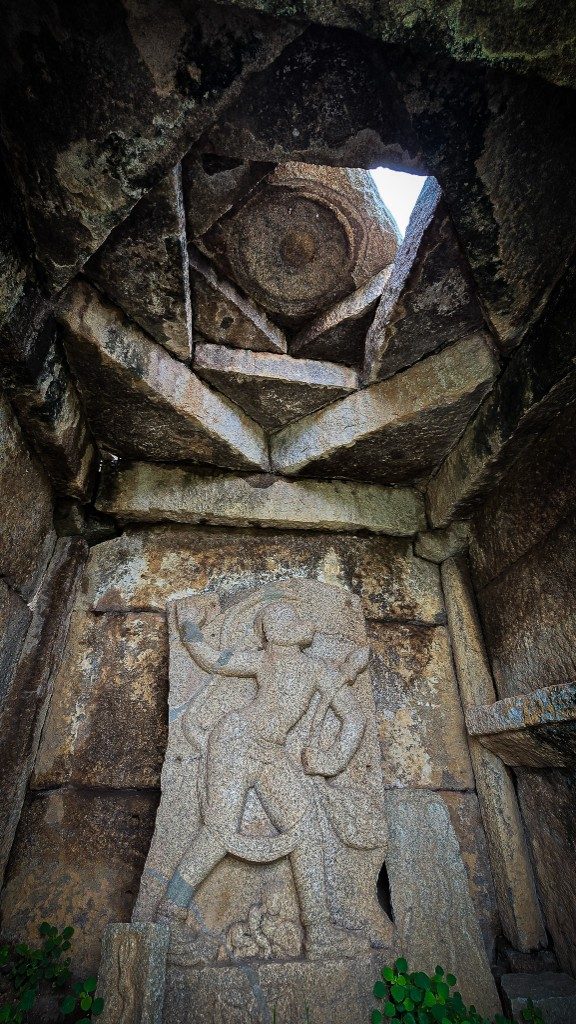 Wide shot of a Hampi stone chamber interior — star-corbelled ceiling above, a carved deity visible in the shadowed base