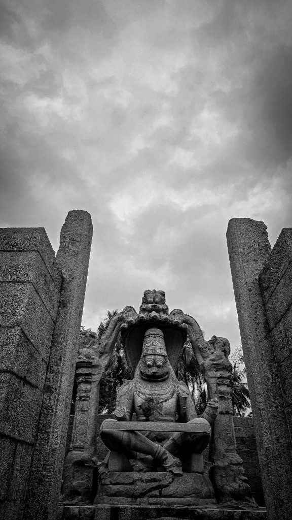 Monumental black and white photograph of the Narasimha sculpture at Hampi — the deity seated under a seven-headed serpent canopy against a heavy clouded sky