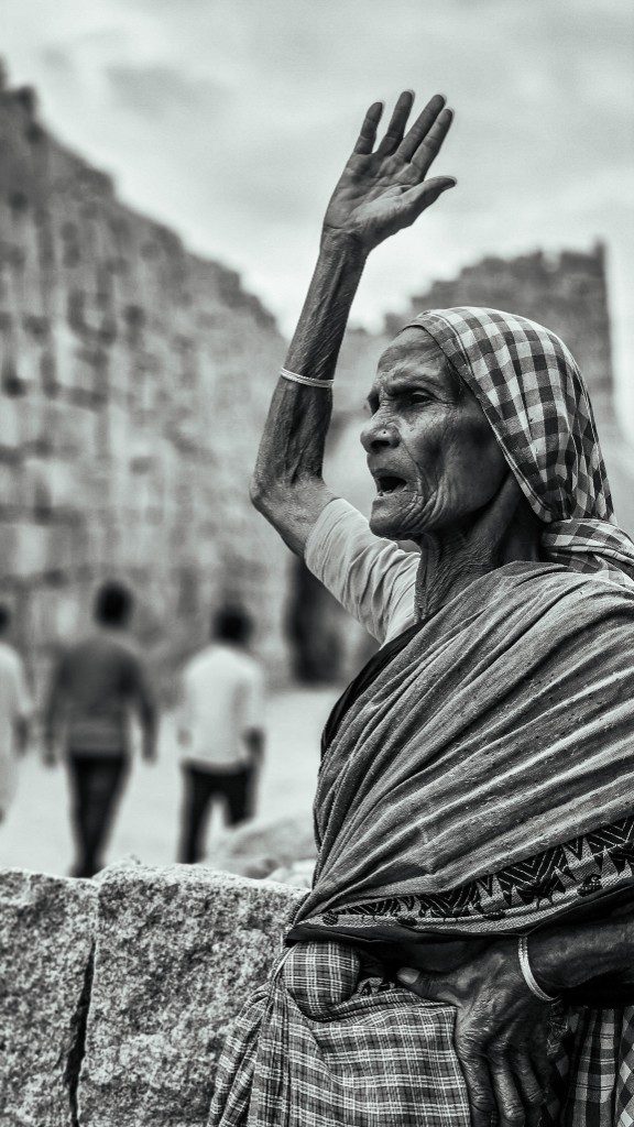 Saratha Amma — an elderly woman in traditional checked saree raises her right hand with authority toward the monuments of Hampi. Her face carries more than forty years of knowledge. The ruins of Hampi are blurred softly behind her.