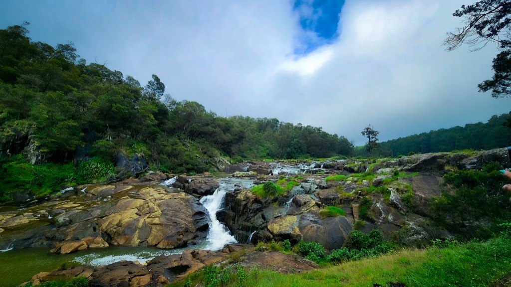 Rocky river cascading over mossy boulders through a green forest clearing under blue sky