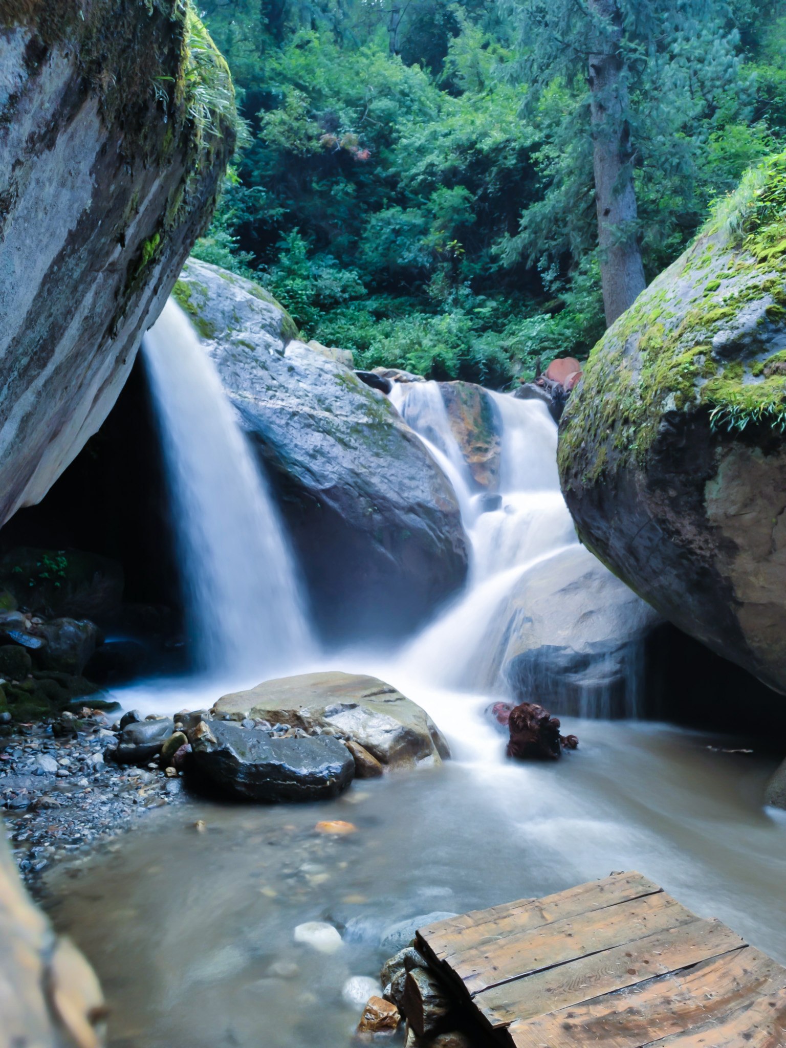 Hidden waterfall tumbling between large mossy rocks in a dark forest with a wooden plank bridge